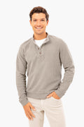 A man smiles, wearing a light gray Marine Layer eco-friendly sweater with a buttoned collar over a white shirt and light-colored pants.