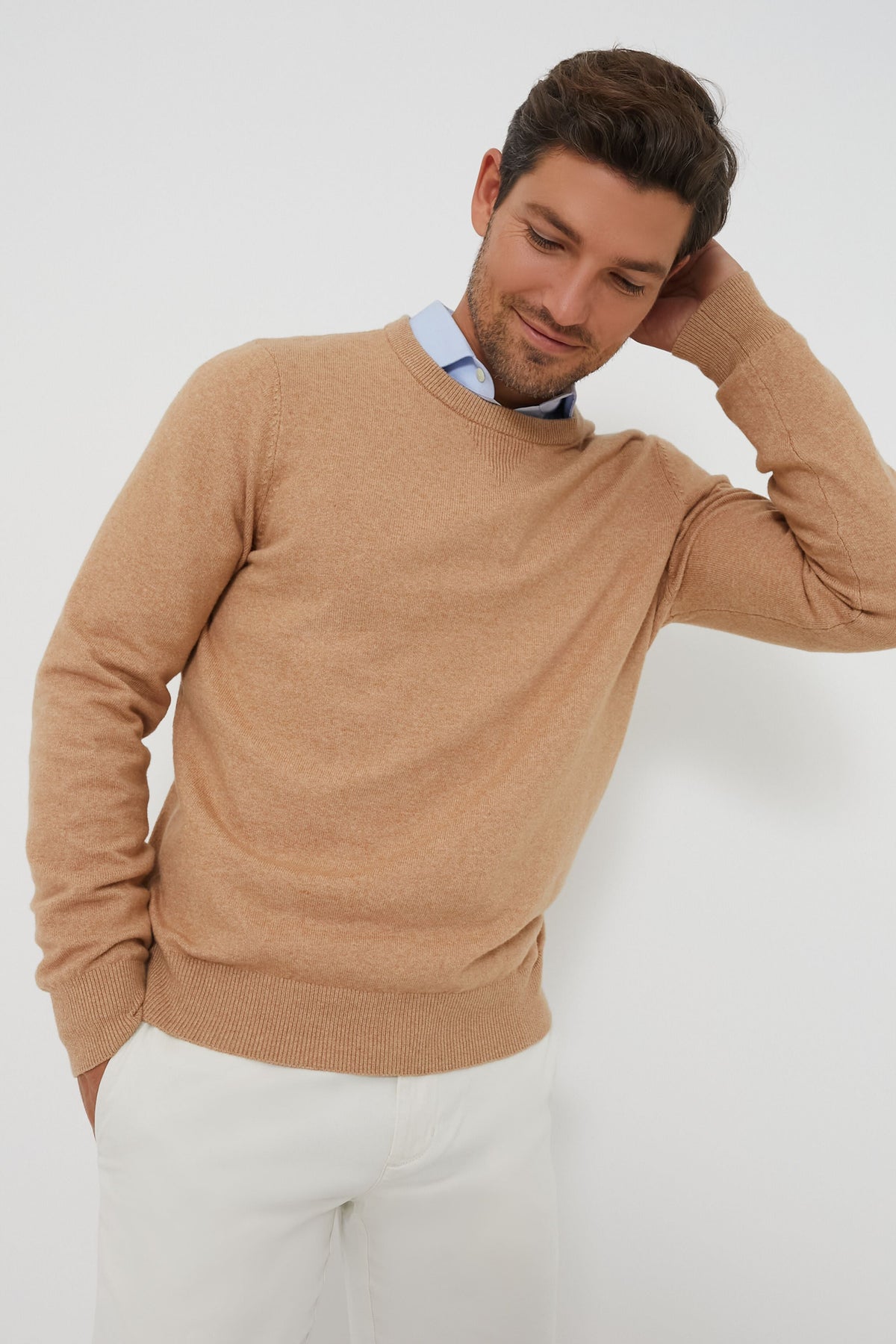 A man in a ORYNESTA sustainable, moisture-wicking crewneck sweater layered over a blue dress shirt and white pants stands confidently, one hand resting on the back of his neck.