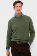 A man stands confidently in a green 100% wool ORYNESTA sweater over a blue collared shirt, paired with brown corduroy pants and a brown belt.