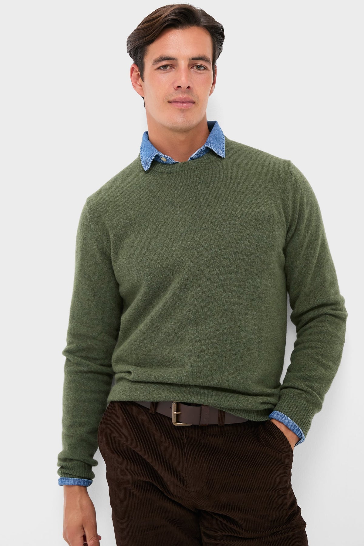 A man stands confidently in a green 100% wool ORYNESTA sweater over a blue collared shirt, paired with brown corduroy pants and a brown belt.